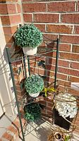 Black metal three-tier plant stand with three faux plants on three shelves, placed against brick wall.