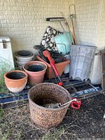 Wide shot showing garden pots including terracotta and plastic, cordless trimmers, garden fencing and cleaning tool handles on a pallet against a white brick wall.
