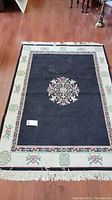 Full view of the rectangular area rug with central black field and cream border with floral accents, shows condition including fringe and surface.