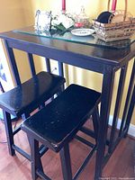 Cafe size black wooden table with glass top and two matching black wooden saddle stools positioned underneath, showing overall look and condition.