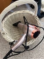 Conair Infiniti Pro blow dryer shown inside a white woven basket on a beige carpet. Device is silver with rose gold accents, black concentrator nozzle attached, black power cord coiled around handle.