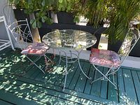 Photo showing round glass-top metal cafe table with two metal chairs on green deck. Chairs have pink patterned cushions and rust visible on metal surfaces.