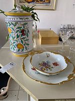 Photo showing Italian ceramic biscotti jar with colorful fruit and leaf designs alongside two French porcelain decorative pieces with floral patterns and gold rims on a white table.