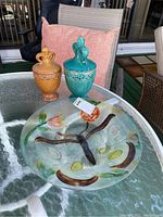 Two ceramic decorative urns (yellow and turquoise) and a round hand-painted glass plate on a glass-top outdoor table with a pink cushion in background.