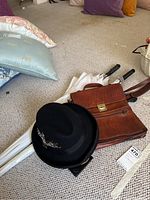 Photo showing black XL fedora hat with decorative feathers, brown leather shoulder bag, and two white umbrellas with black handles.