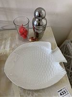 White ceramic fish plate and stainless steel cocktail shaker with clear glass containing red pour spouts on beige cloth surface