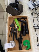 Wooden tray with assorted black pocket flashlights, green utility knife, black and orange pruners, and green tape dispenser.