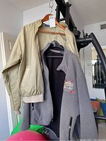 Two jackets hanging on hangers: beige bomber jacket and gray zip-up jacket with logo