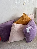 Pile of five oversized square plush pillows in purple, mustard yellow, and pink shades lying on carpet against a wall.