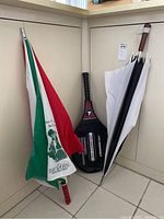 Photo showing two folded sport umbrellas (one tri-color with The Doral Hotels of Florida logo, one black and white) and a Prokennex Celebrity 710 tennis racket standing against a wall.