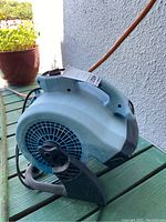 Side view of light blue X Power air mover showing air vent and black molded stand base on green wooden surface with plant background.