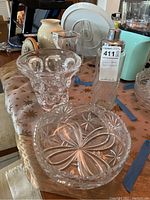 Photo showing three crystal glass items: a textured glass vase, a starburst patterned bowl, and a tall etched glass shaker with metal top, all placed on a table.