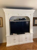 Front view of the tall white Baker bookcase with TV in the middle, built-in upper shelf holding electronic devices, and closed base cabinets with brass knobs.