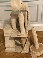 Side view of beige plush cat tree showing multiple levels, two vertical scratching posts (one detached), and a hanging ball toy attached with a rainbow-striped ribbon.
