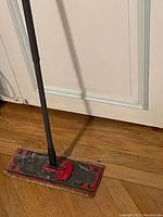 Photo showing the floor mop with a rectangular black and red cleaning head and black handle, placed against a wall on a wooden floor.