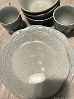 Close-up photo of Lenox French Pearle dinnerware set showing plate in center foreground with three stacked bowls behind and three mugs on each side, all featuring the textured and embossed decorative pattern typical of the French Pearle design.