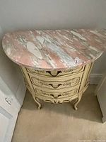 Front view of the cream wooden side table with pink and white marble top showing three curved drawers and decorative carved legs.
