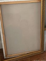 Front view of an empty rectangular picture frame with a gold-colored ornate border and wooden backing.