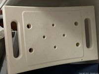 Photo shows white portable seat made of molded plastic with two handles and multiple round ventilation holes on surface.