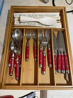 Top down photo of a wooden cutlery tray holding 4 spoons, 4 forks, and 6 knives with red handles along with some wrapped chopsticks and a white spoon.