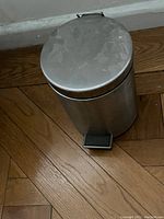 View of single small stainless steel pedal trash can with lid and foot pedal, shows surface marks and dirt on top.