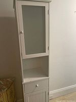 Tall white wooden cabinet with a frosted glass door on top, an open shelf below it, a drawer under the shelf, and a solid lower door. Cabinet has round white knobs on each section and is placed against a wall.