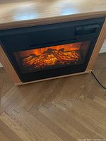 Virtual sizzling electric fireplace insert inside a wooden console, showing glowing ember and flame effects, plugged in with power cord visible.