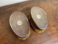 Pair of oval sterling silver brushes with natural horse hair bristles shown from above on a wooden surface, featuring monogram plaques.