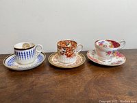 Three antique demitasse cups and saucers shown side by side on a wood surface, showcasing different floral and geometric designs.