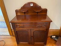 Front view of antique mahogany washstand showing carved backsplash with two circular platforms, drawer with moustache-shaped pulls, and double cabinet doors.