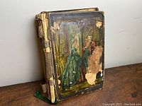 Front view of antique Victorian scrapbook album showing worn fabric spine, damaged covers depicting Victorian figures in period dress, and some areas of loss.