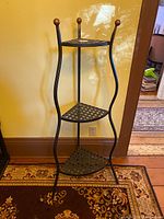 Full view of the 3-tier wrought iron corner shelf standing on a patterned rug against a yellow wall, showing curved vertical metal supports and triangular lattice shelves.