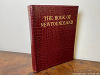 Image of the hardcover book showing the textured maroon cover with gold embossed title "THE BOOK OF NEWFOUNDLAND".