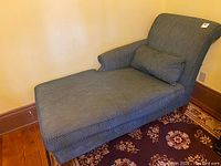 Full view of the upholstered fainting couch showing the curved backrest, rolled armrest, and bolster pillow on textured greenish-grey upholstery.