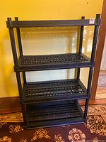 Full view of the assembled four-tier black plastic shelving unit against a yellow wall, showing the top and three additional shelves.