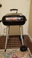 Full view of the black 17.5 inch charcoal BBQ grill showing legs, wheels, storage rack, and lid with handle