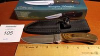 Cougar Hunter hunting knife with polished steel blade shown next to a wooden ruler indicating about 8 inches overall length; black fabric sheath below knife; product box in background with knife image and branding.