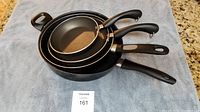 Four stacked black Faberware cooking pans with handles visible, laid on a grey cloth.