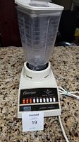 Full view of Osterizer blender with clear pitcher and white plastic lid on granite countertop.