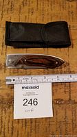 Photo of pocket folding knife with wooden handle and stainless steel blade, measuring about 8 inches overall, placed next to black fabric sheath and ruler on wooden surface.