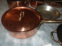 Copper cookware items arranged on a soft surface for condition and detail photographing. Shows details of copper exterior and brass handles.