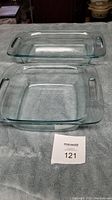 Two clear glass Pyrex cooking pans, one square approximately 8x8 inches, one rectangular approximately 10x7 inches, both with handle slots on sides.