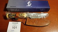 Knife with protective plastic on the blade, bone-style handle, and leather sheath with embossed logo. Box in background shows brand and specifications.