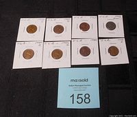 Top-down image showing 10 US pennies in white cardboard holders arranged in two rows on dark fabric background with auction lot number card visible.