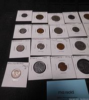 Photo of 16 coins from various countries in white cardboard holders with handwritten country and year labels.