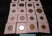 Coins in cardboard holders arranged in rows with dates and country names handwritten on the holders.