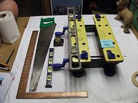 All carpentry tools arranged on white surface, showing table top workmate, handsaw, levels, rafter square, tape measure grouped for lot view.