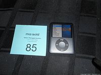 Front view of the Apple iPod Nano showing the color screen and click wheel, with a MaxSold seller auction card showing number 85