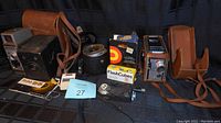 Full lot of vintage camera equipment including Bell and Howell video camera, Kodak box camera, light meters, flash cubes, and flash arm with diffuser arranged on fabric background.