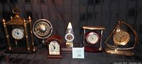 Six battery operated clocks displayed on a black fabric background showing varying styles including brass, wood and clear plastic pyramid case.